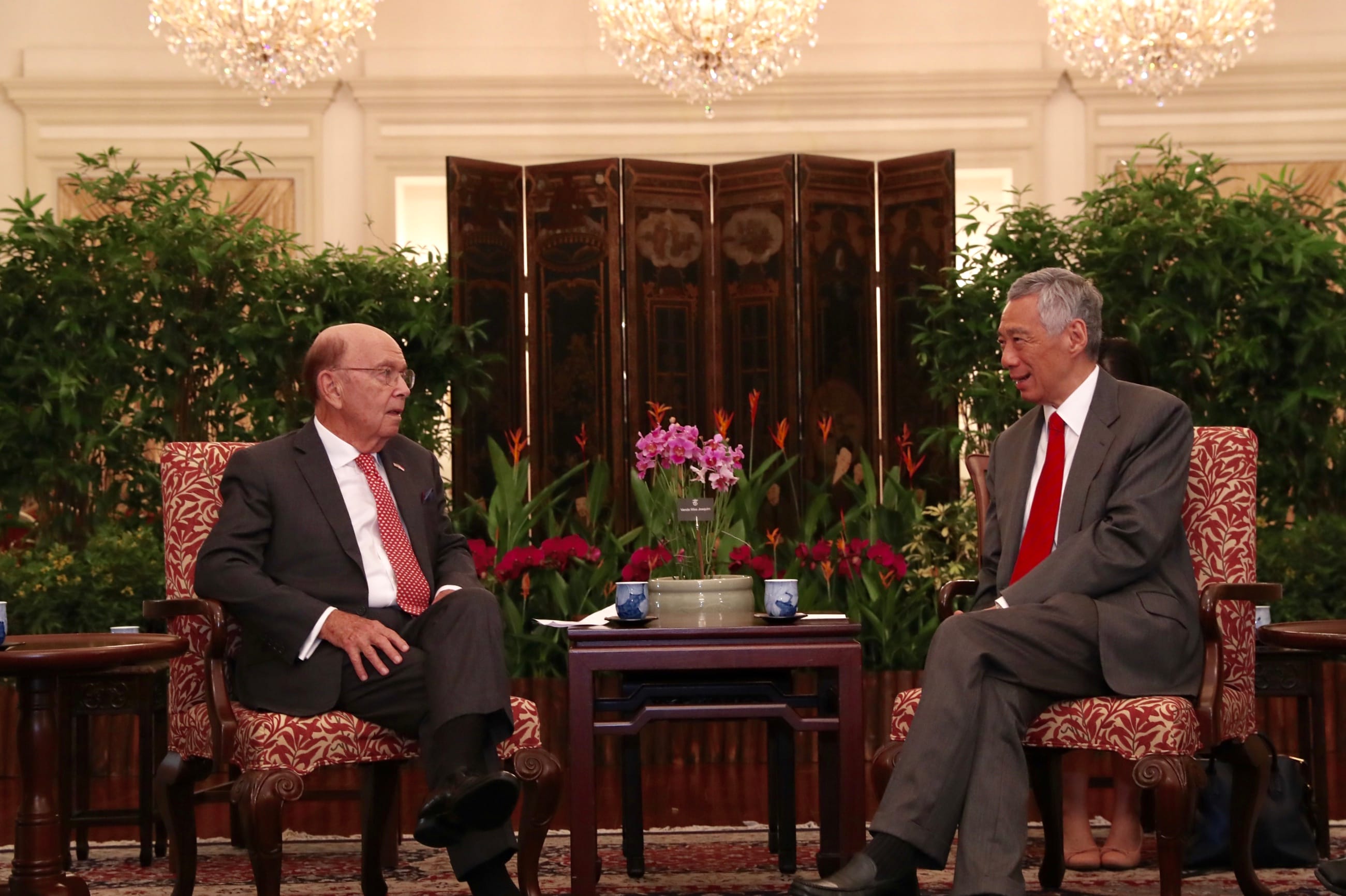 Wilbur Ross and Lee Hsien Loong seated in patterned chairs, facing each other indoors. A small table separates them.